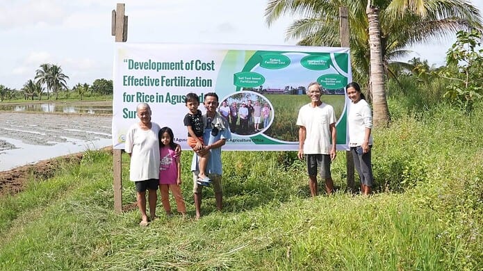 TESTING BEFORE PLANTING. Rice-farming families in Agusan del Sur stand before a project billboard for the "Development of Cost Effective Fertilization for Rice" program. Rep. Adolph Edward "Eddiebong" Plaza cited the pilot initiative as proof that science-based soil diagnostics can help smallholders slash input costs and improve harvest yields by doing away with guesswork. Photo courtesy of Team Eddiebong page